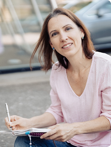 Portrait von Simone Bürger, Grafikdesignerin aus Wernau, sitzend mit einem Notizbuch in der Hand, Blick in die Kamera, rosa Bluse und Jeans