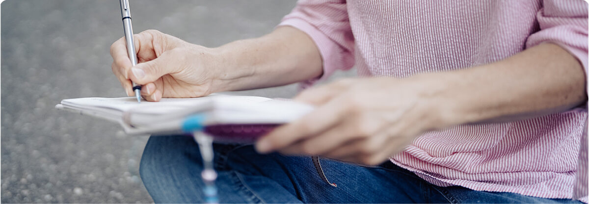 Simone Bürger schreibt etwas in ihr Notizbuch, sitzend mit rosa Bluse und Jeans